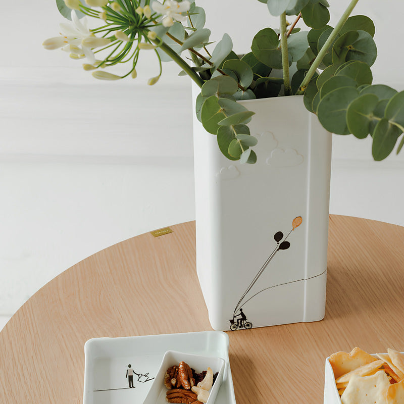 Aus der Bildergeschichten-Kollektion von Räder Design entstammt diese Blumenvase mit dem Namen REGENWOLKE. Unser Radfahrer hat es eilig, seine Ballons fliegen an der Außenseite der Vase in den Himmel. Sie ist aus weißem Porzellan, hat eine quadratische Form und eine Größe von 12x12x25 cm und ist somit für Blumen mit Stiel für mittlere Länge geeignet.